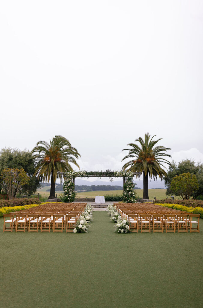 Outdoor wedding ceremony setup with rows of wooden chairs, white floral arrangements lining the aisle, and two palm trees framing an arbor on a lawn.
