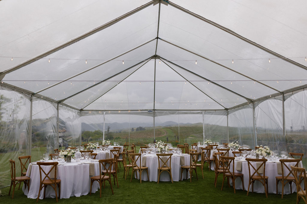 A clear tent set up outdoors with round tables covered in white tablecloths, wooden chairs, and floral centerpieces, ready for an event.