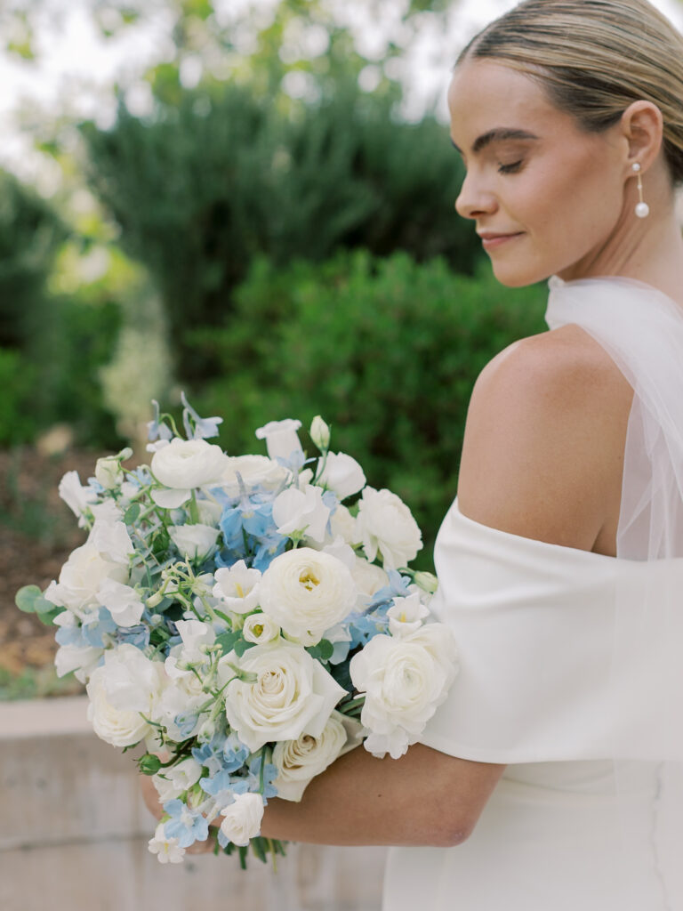 A bride in an off-shoulder white dress holds a bouquet of white and light blue flowers, standing outdoors among lush greenery at a beautiful Solage Calistoga wedding.