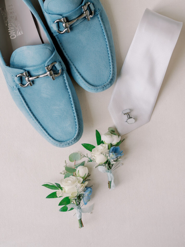 Light blue loafers, a white necktie with cufflinks, and two small boutonnieres with white and blue flowers arranged on a white surface—perfect details for a Solage Calistoga wedding.