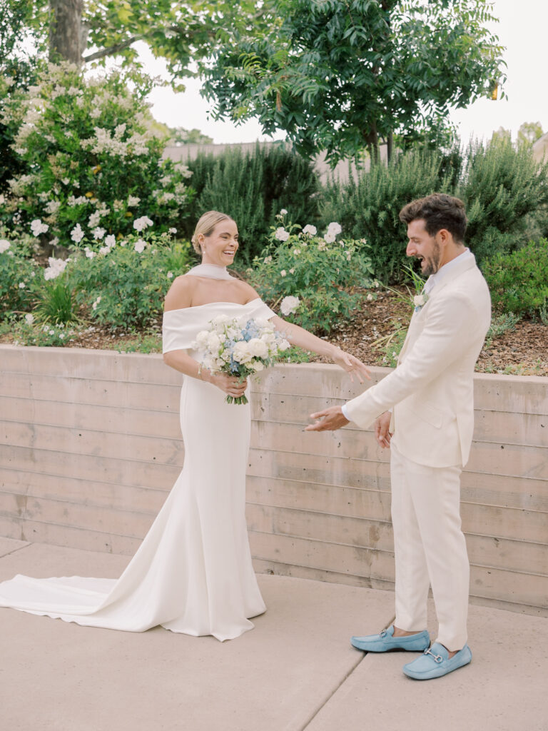 A bride in an off-shoulder white gown holds a bouquet and smiles at the groom in a white suit and blue shoes, standing outdoors near greenery at their Solage Calistoga wedding.