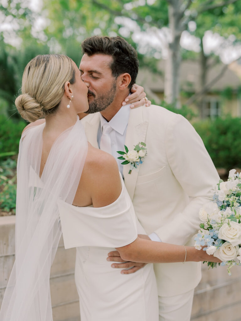 A bride and groom dressed in white kiss outdoors at their Solage Calistoga wedding; the bride holds a bouquet of white flowers and greenery.