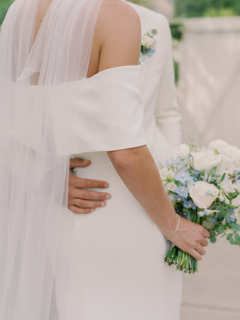 A bride in a white dress holds a bouquet of white and blue flowers while standing with her partner at their Solage Calistoga wedding, his hand gently resting on her waist.