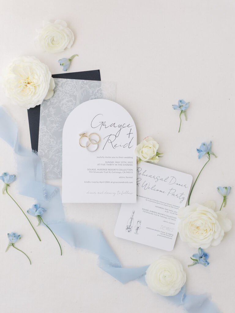 Elegant Solage Calistoga wedding invitation suite featuring white and translucent cards, gold rings, blue ribbon, and white and blue flowers beautifully arranged on a light surface.