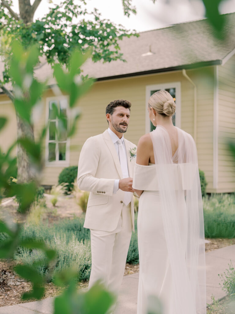 A bride and groom in white outfits stand facing each other outside by a yellow house, surrounded by greenery at their Solage Calistoga wedding.