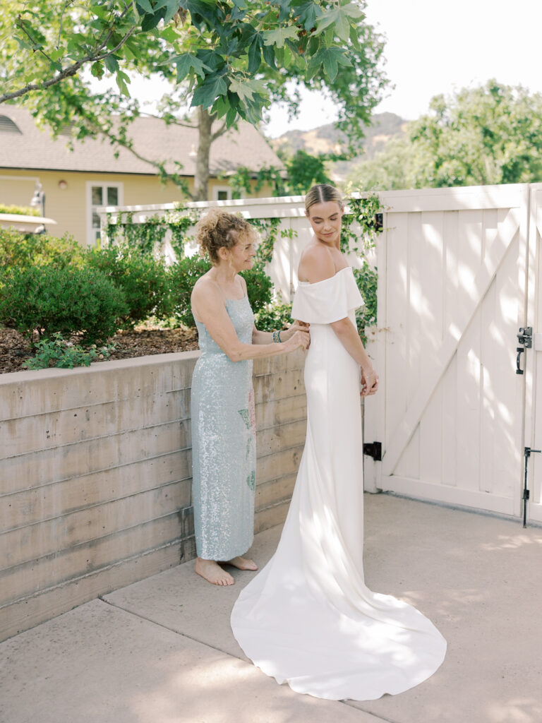 A woman in a light blue dress helps a bride in an off-shoulder white gown adjust her dress outdoors by a white fence on a sunny Solage Calistoga wedding day.