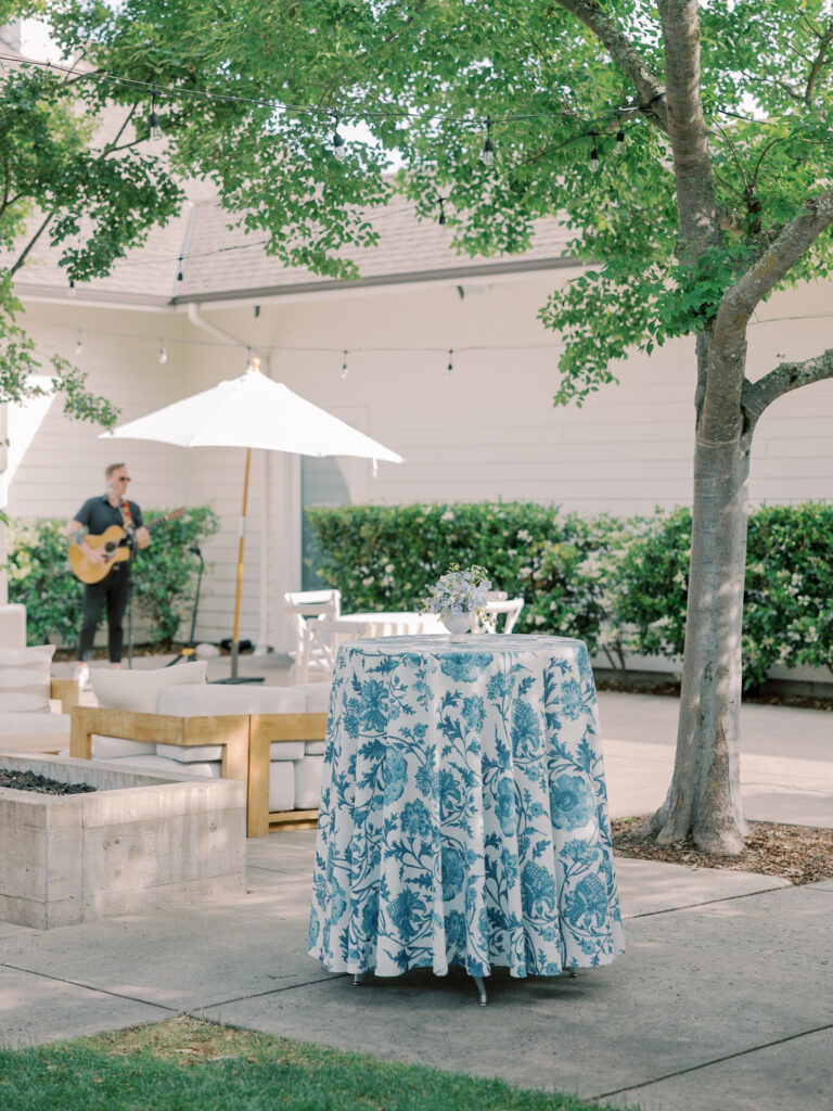 A round table with a blue floral tablecloth stands on a patio; nearby, a man plays guitar under a white umbrella, evoking the relaxed elegance of a Solage Calistoga wedding, surrounded by trees and outdoor seating.