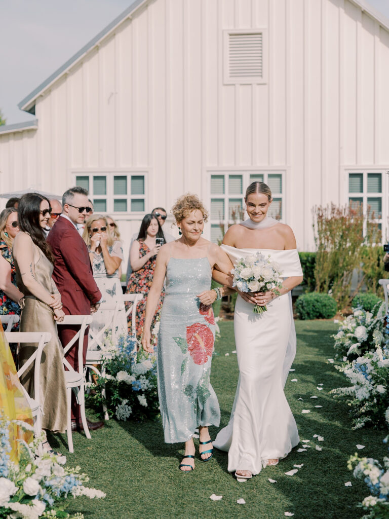 A bride in a white dress walks down an outdoor aisle with an older woman in a floral dress, surrounded by guests at a stunning Solage Calistoga wedding in front of a beautiful white building.