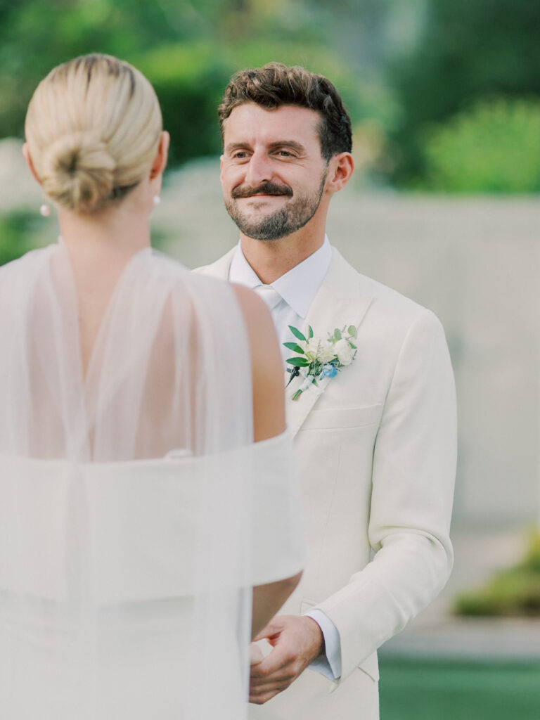 A man in a white suit with a boutonniere smiles at a woman in a white dress and veil during an outdoor Solage Calistoga wedding ceremony.