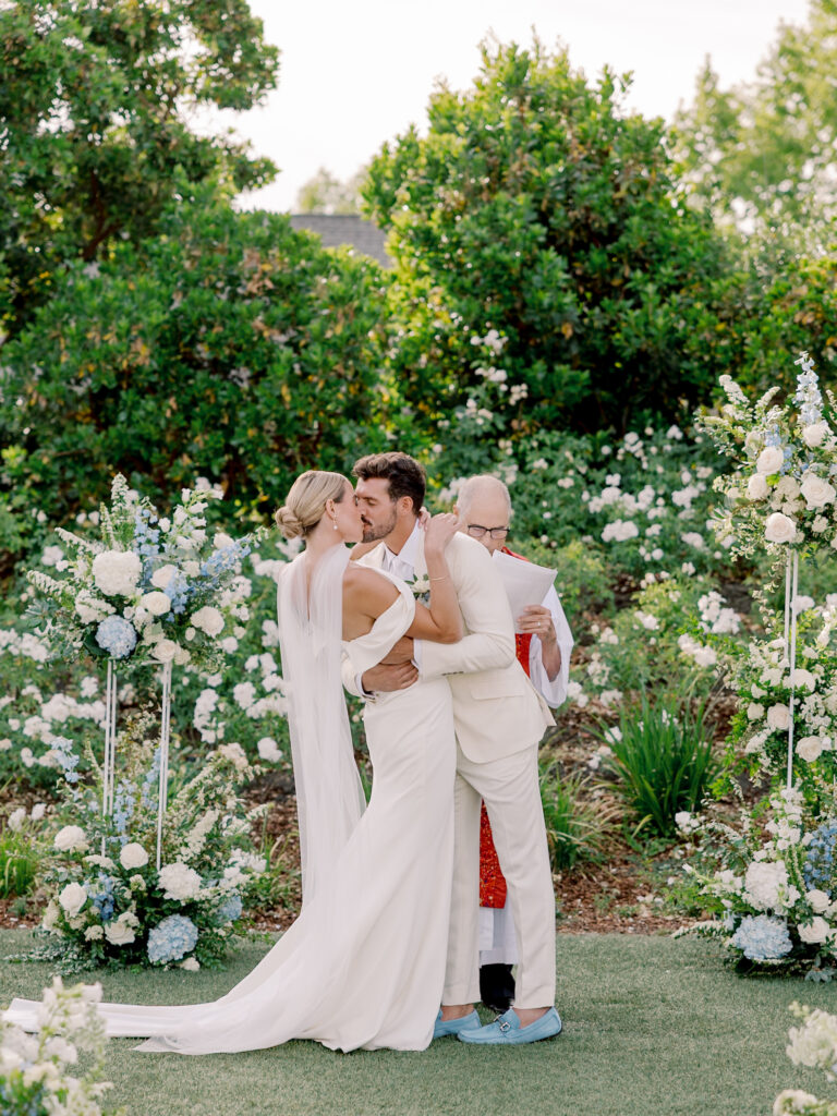 A bride and groom kiss at their Solage Calistoga wedding ceremony, surrounded by white and blue flowers, while an officiant stands behind them holding papers.