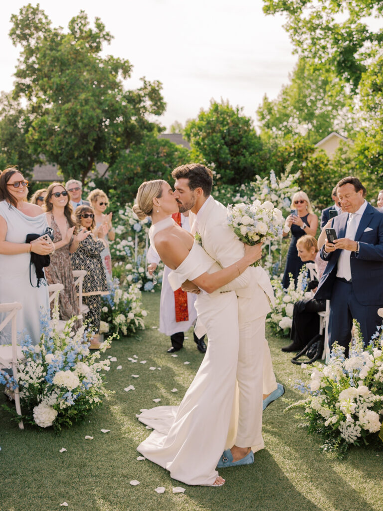 A newlywed couple kisses while walking down an outdoor aisle lined with flowers at their Solage Calistoga wedding, surrounded by applauding guests capturing the moment.