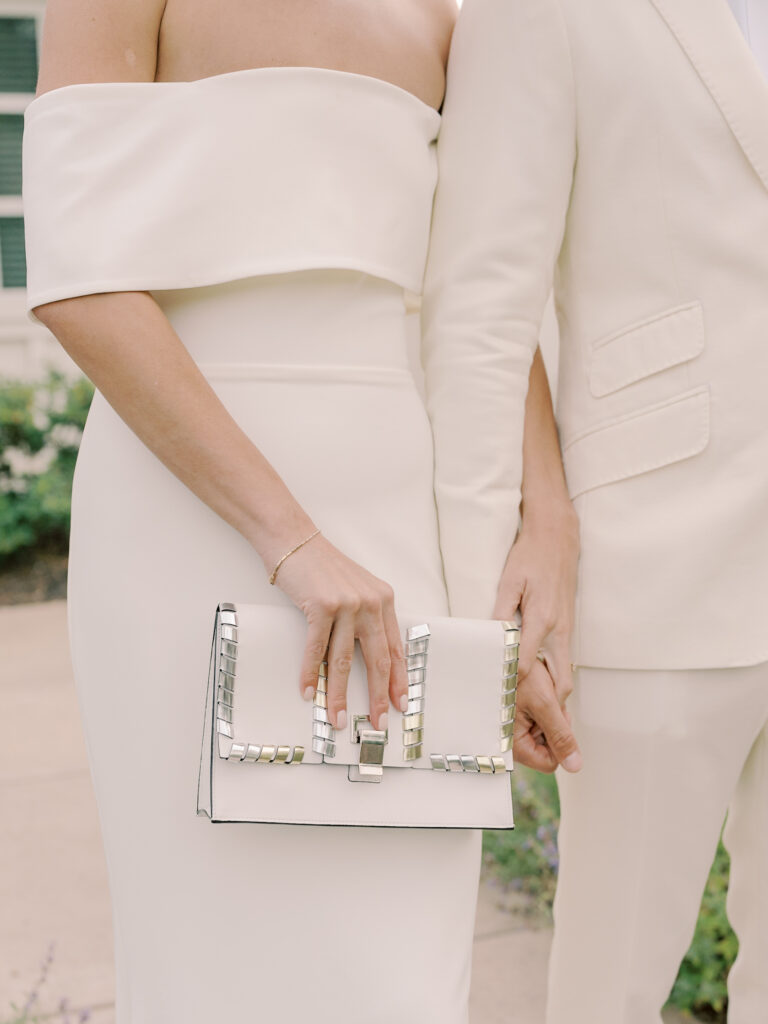 Two people dressed in elegant, off-white formal attire stand side by side at a Solage Calistoga wedding, one holding a white clutch with metallic accents.
