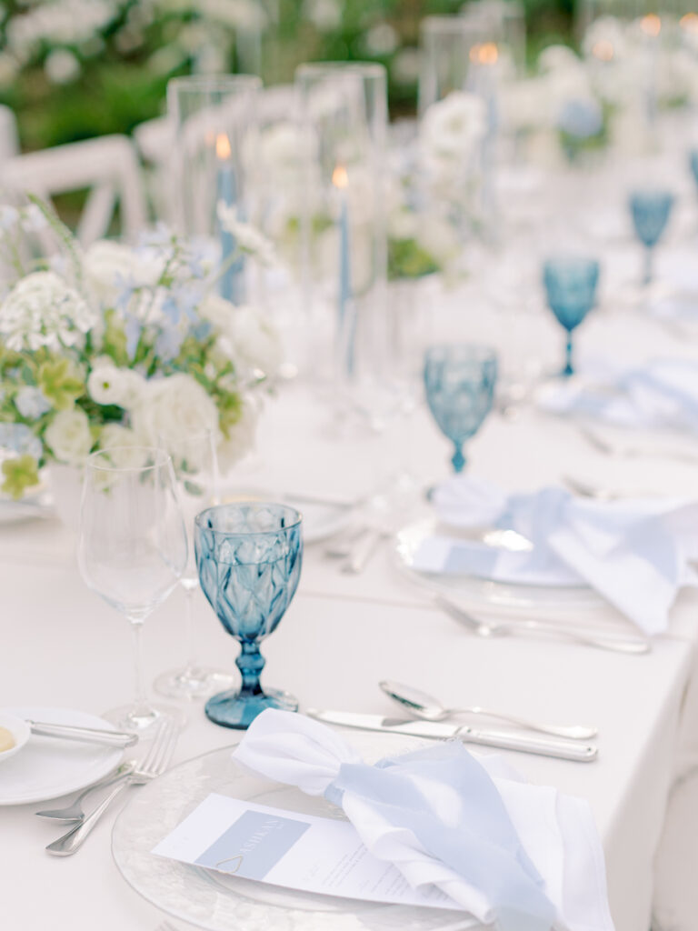Elegant outdoor table setting at a Solage Calistoga wedding with white linens, blue glass goblets, white floral arrangements, candles in glass holders, and neatly folded napkins on clear plates.