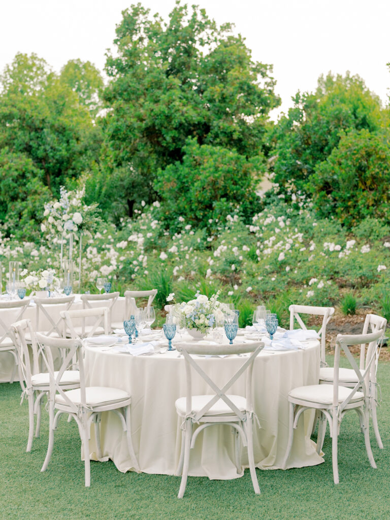 Round table covered with a white tablecloth, set with blue glassware and white floral centerpieces, surrounded by white chairs in an outdoor garden setting perfect for a Solage Calistoga wedding.