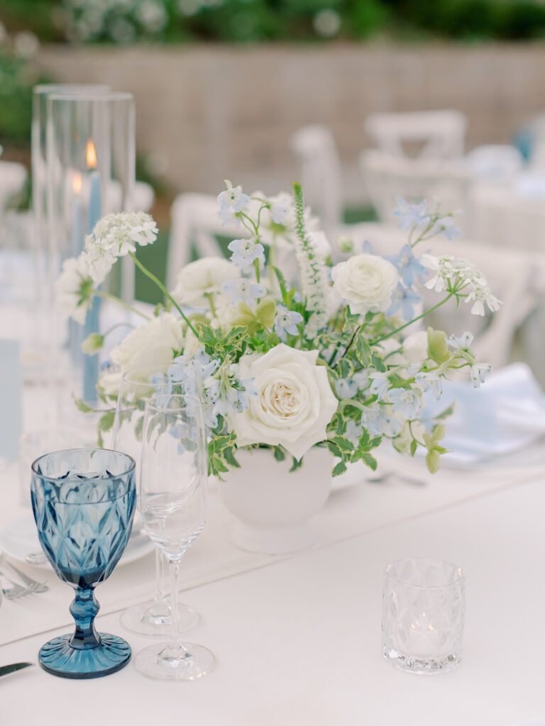Elegant table setting at a Solage Calistoga wedding with a white floral centerpiece, blue glass goblet, clear wine and water glasses, a votive candle, and a tall glass candle holder atop a crisp white tablecloth.