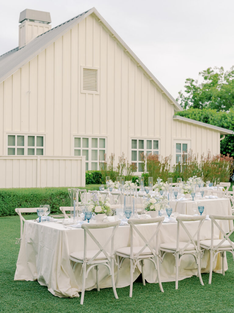 Two long tables with white tablecloths and blue glassware are set up outdoors on grass in front of a cream-colored building, capturing the elegant charm of a Solage Calistoga wedding.