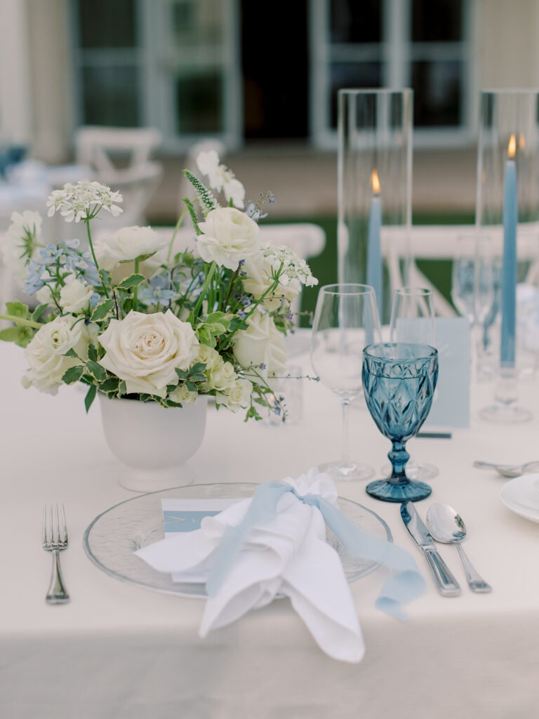 Table setting at a Solage Calistoga wedding with white flowers in a vase, blue glass goblet, clear plate, blue and white napkin, candles, and silverware on a white tablecloth.