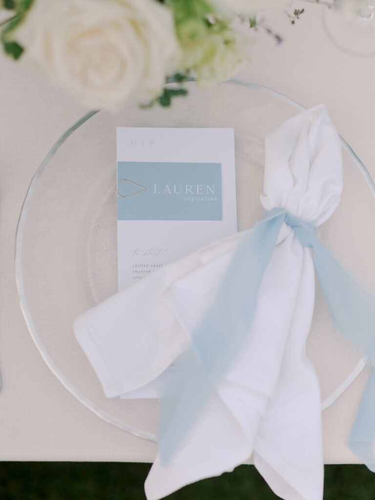 A clear glass plate with a folded white napkin tied with a blue ribbon and a blue and white menu card labeled "LAUREN" rests on a cream tablecloth, capturing the elegant style of a Solage Calistoga wedding.