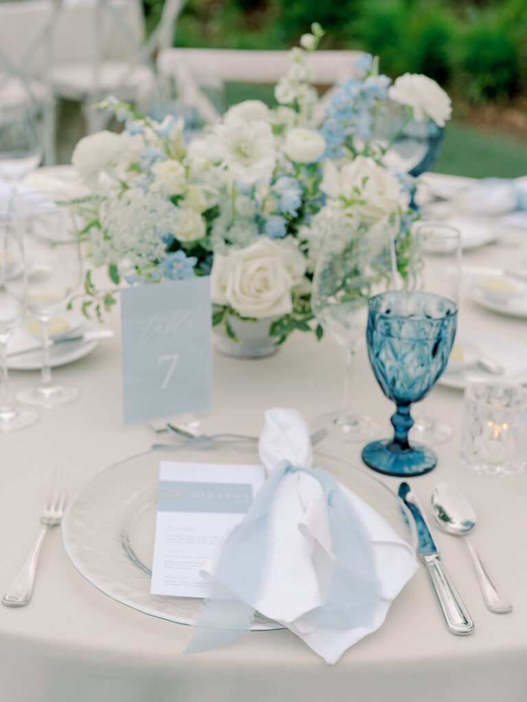 Table setting with a clear plate, menu, white napkin, blue glass, and silver cutlery. A floral centerpiece with white and blue flowers and a "Table 7" card evoke the elegance of a Solage Calistoga wedding.