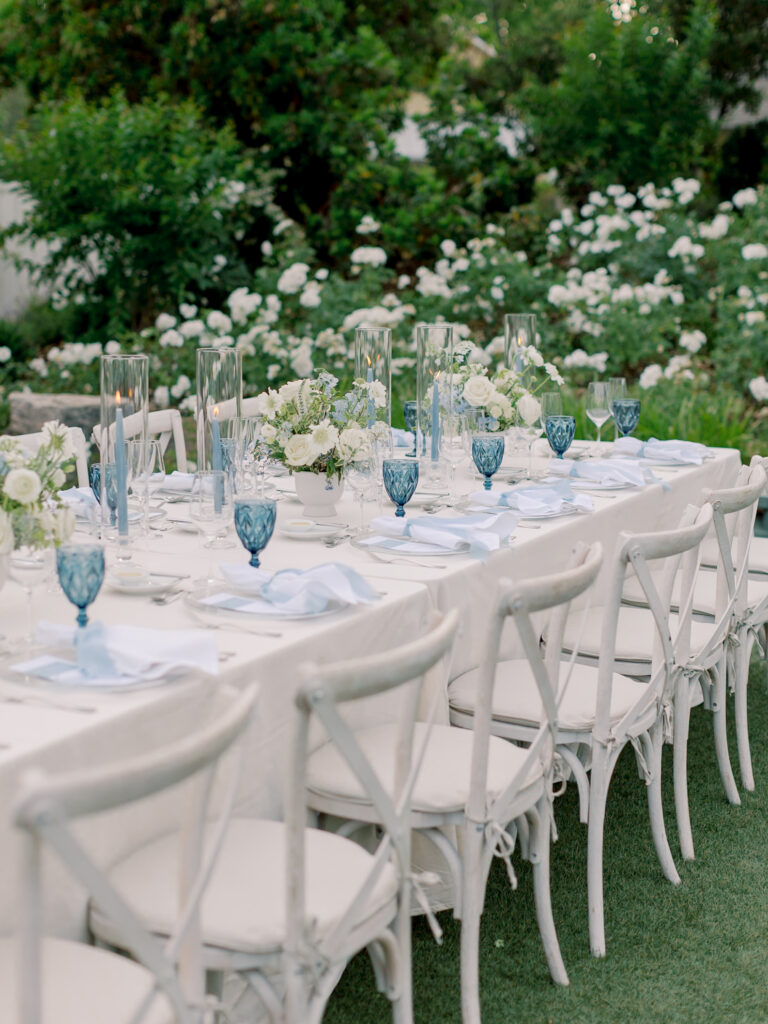 A long outdoor dining table with white chairs, blue glassware, white plates, and tall glass candle holders set in a garden—perfect for an elegant Solage Calistoga wedding celebration.