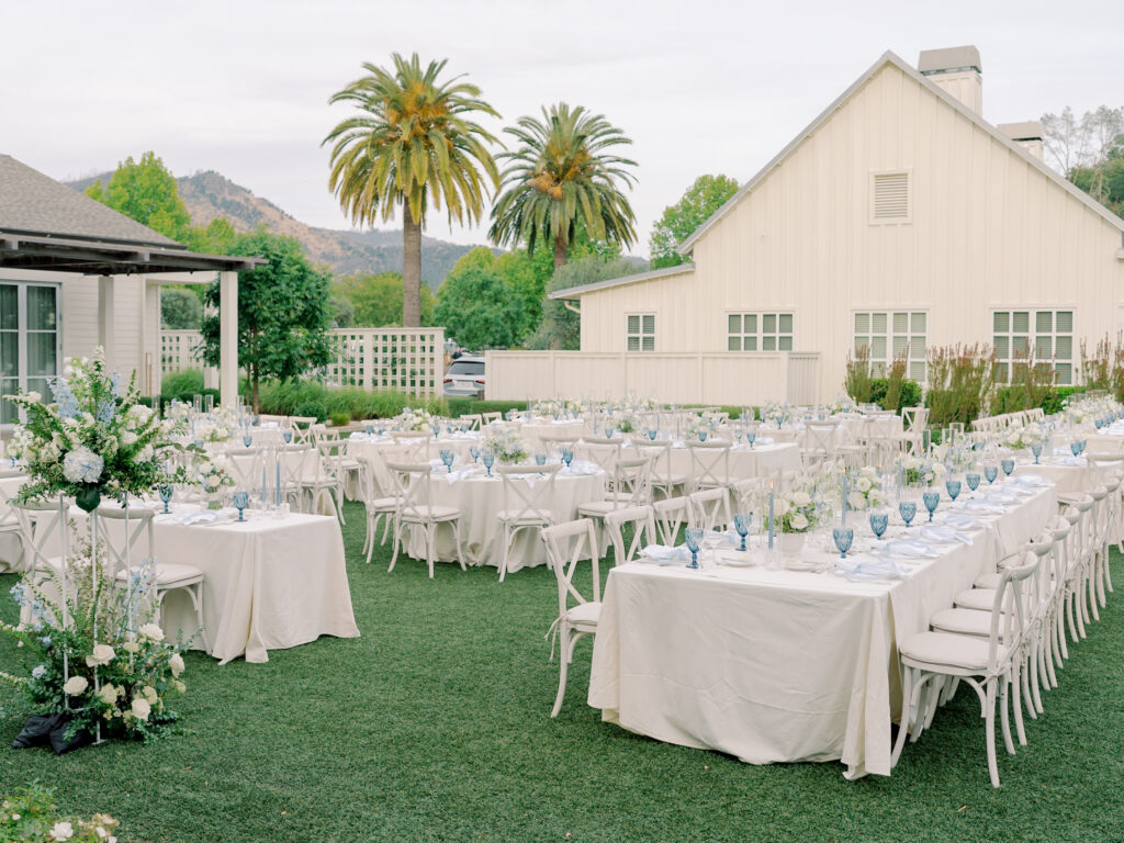 Outdoor Solage Calistoga wedding reception setup with long tables covered in white linens, blue glassware, and floral arrangements, set on grass near a white barn and palm trees.