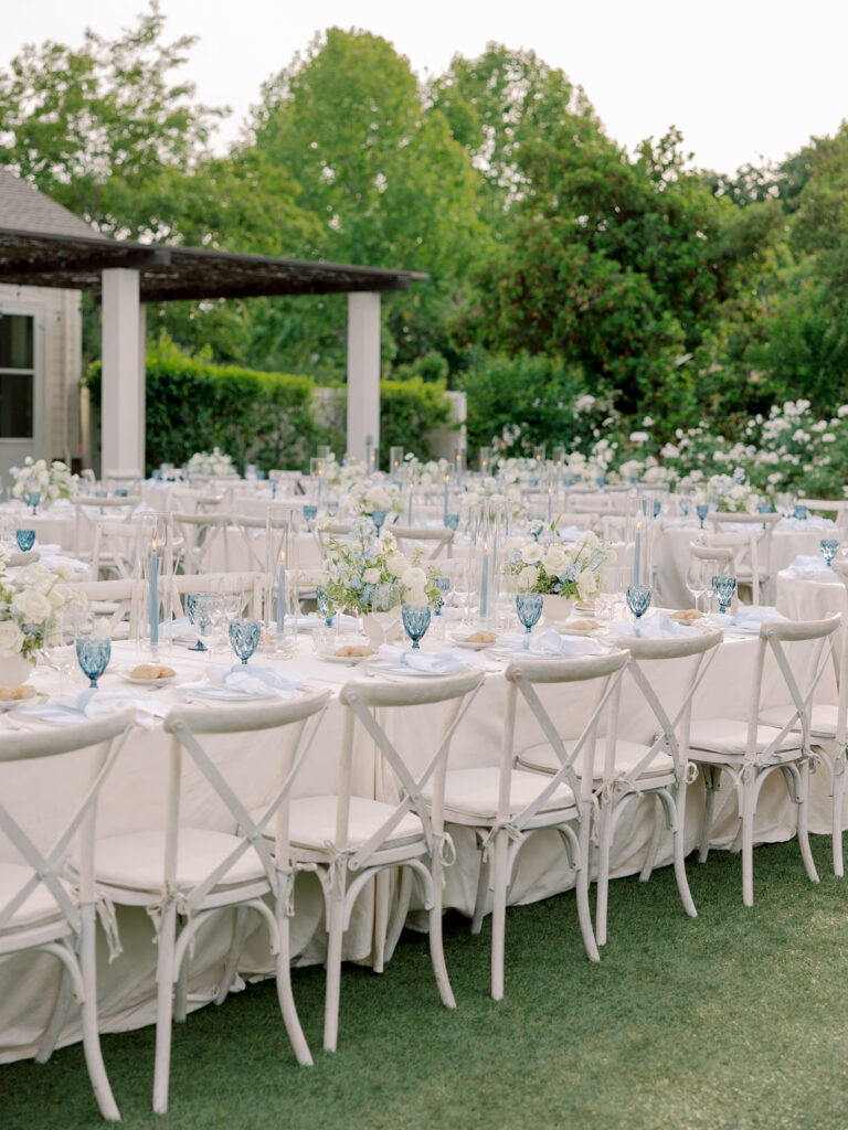 Long tables with white tablecloths, white chairs, blue glassware, and white floral centerpieces are arranged outdoors on grass, surrounded by greenery at a beautiful Solage Calistoga wedding.