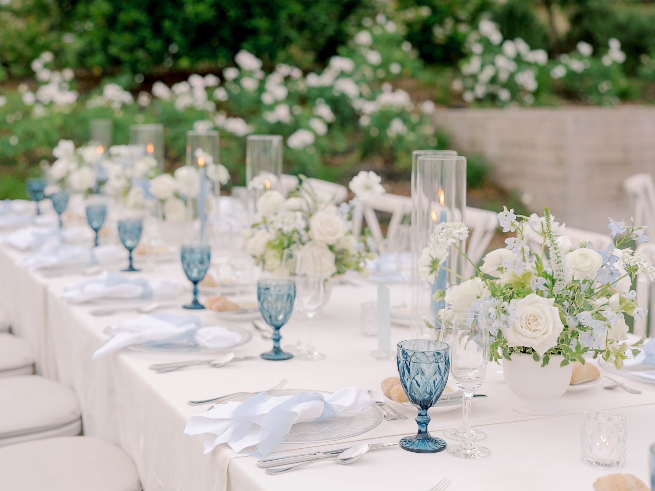 An elegant Solage Calistoga wedding outdoor table setting featuring white linens, blue glassware, white floral centerpieces, lit candles in tall holders, and neatly arranged cutlery and napkins.