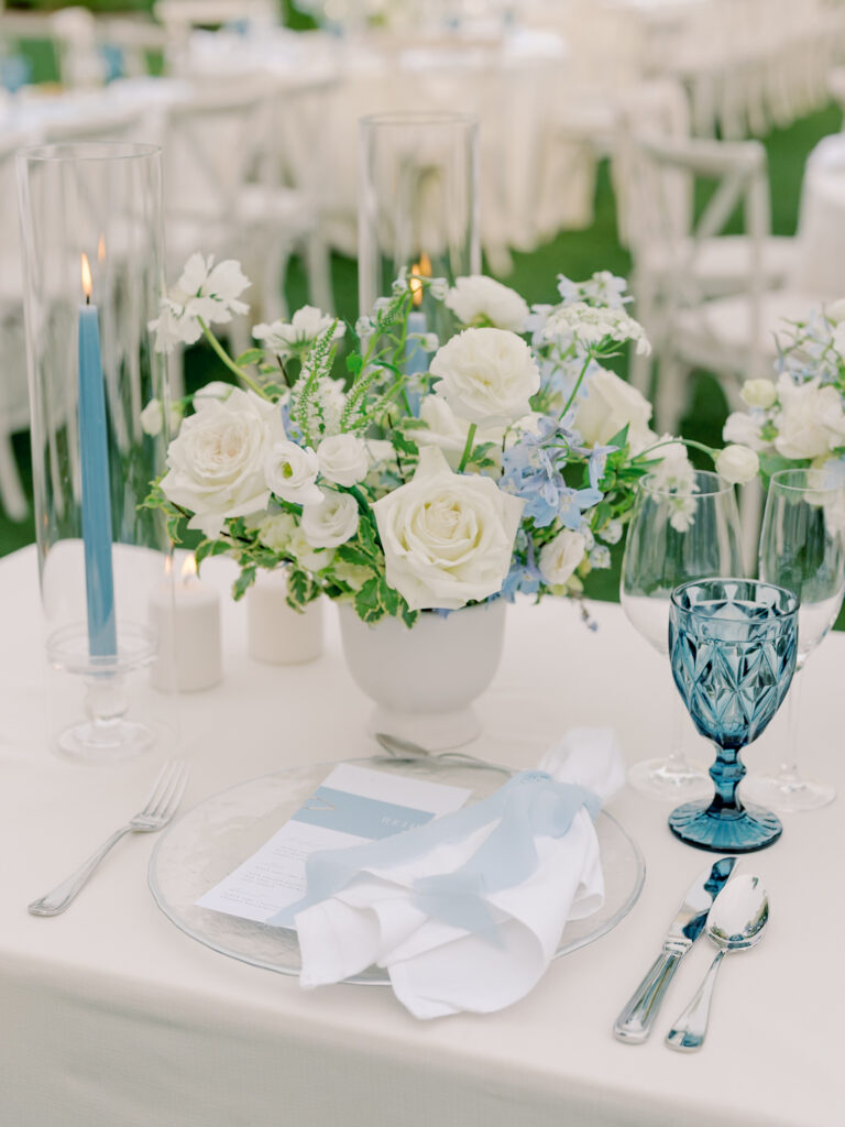 Elegant table setting at a Solage Calistoga wedding with a white and blue floral centerpiece, blue candle, blue glass goblet, white napkin, and menu on a clear plate.