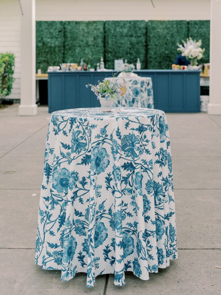 A round table with a blue floral tablecloth and a small flower arrangement stands outdoors at a Solage Calistoga wedding; a bar setup is visible in the background.