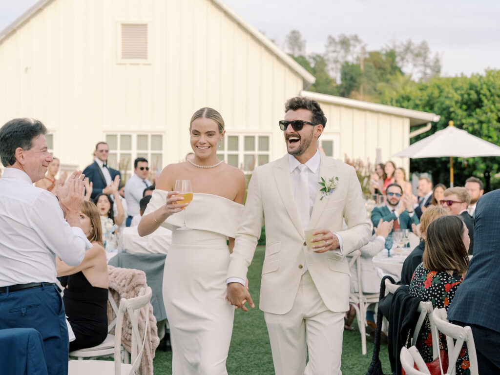 A bride and groom in white attire walk hand in hand outdoors at their Solage Calistoga wedding, smiling and holding drinks, as wedding guests seated at tables applaud.
