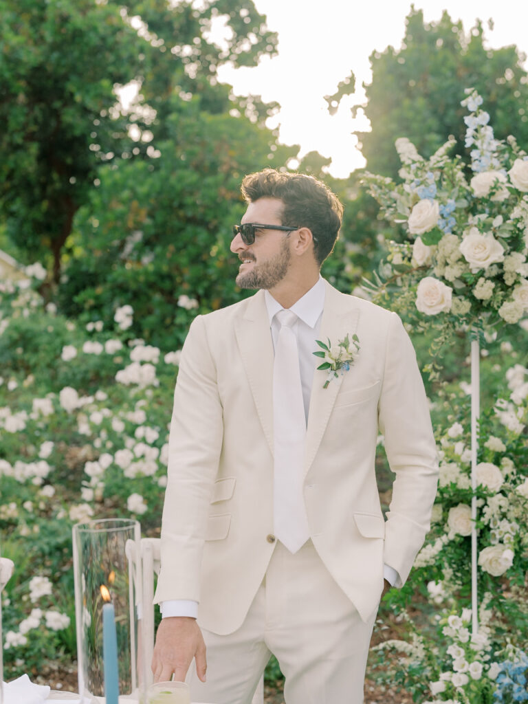 A man in a cream suit and sunglasses stands outdoors near a floral arrangement, surrounded by white roses and greenery at a Solage Calistoga wedding.