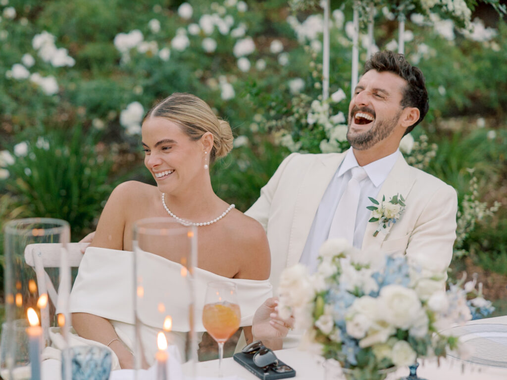 A bride and groom sit together at a decorated table outdoors at their Solage Calistoga wedding, smiling and laughing, surrounded by flowers and candles.