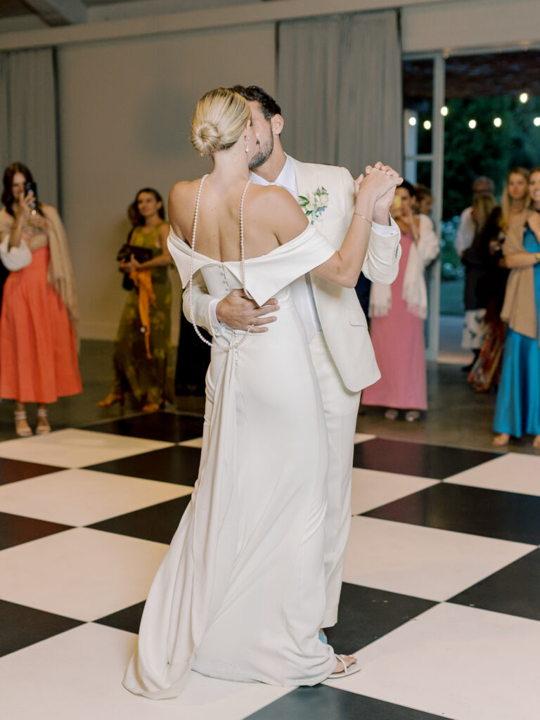 A bride and groom in white attire share a dance on a black and white checkered floor at their Solage Calistoga wedding while guests watch in the background.