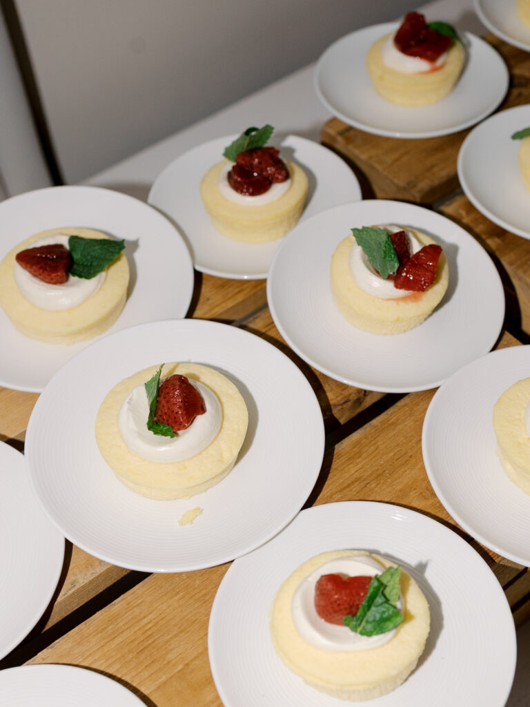 Small sponge cakes topped with whipped cream, a strawberry half, and a mint leaf are arranged on white plates over a wooden surface—perfect for a Solage Calistoga wedding dessert display.