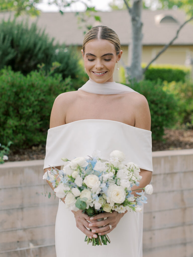 A woman in an off-shoulder white dress holds a bouquet of white and light blue flowers, standing outdoors by lush greenery and a low stone wall at a Solage Calistoga wedding.