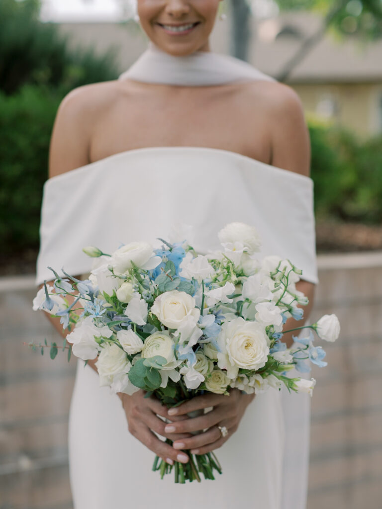 A woman in an off-the-shoulder white dress holds a bouquet of white and light blue flowers, standing outdoors with lush greenery—a dreamy Solage Calistoga wedding scene.