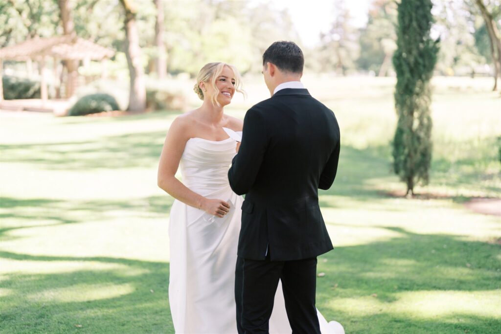 A bride and groom stand facing each other outdoors on a sunny day; the bride is smiling and holding a piece of paper, while the groom wears a black suit.