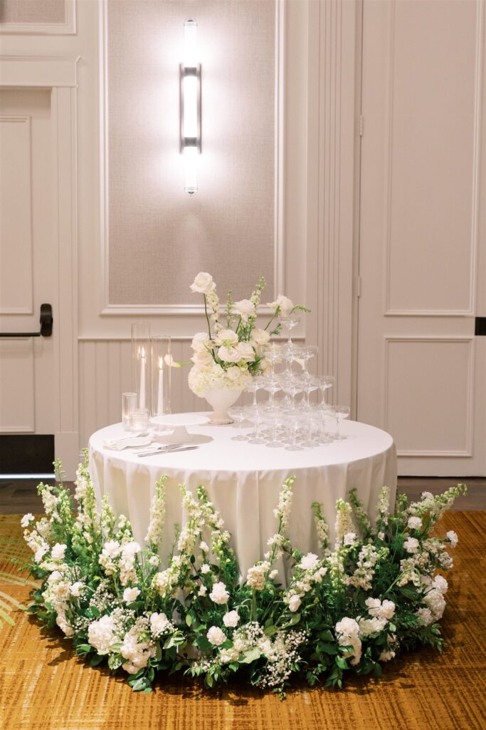 Round table with white tablecloth, surrounded by white and green floral arrangements, holds stacked champagne glasses, candles, and a white flower centerpiece in an elegant indoor setting.