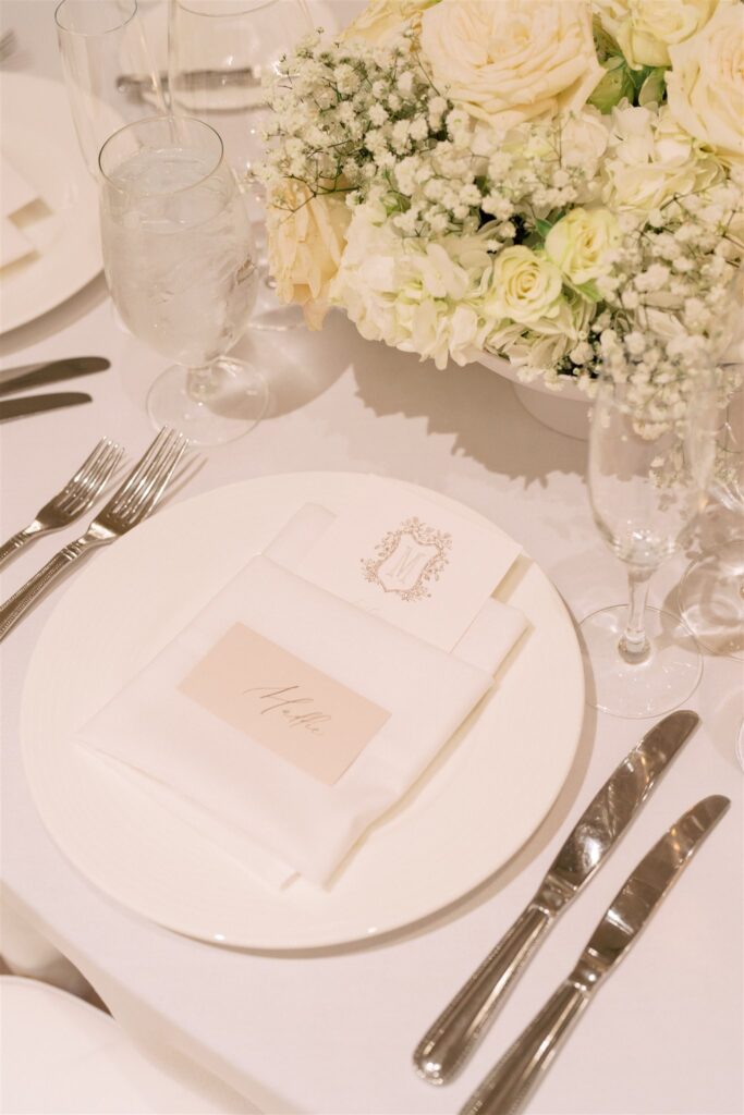 Elegant table setting with white plates, silver cutlery, a folded napkin with a name card, water and champagne glasses, and a centerpiece of white flowers.