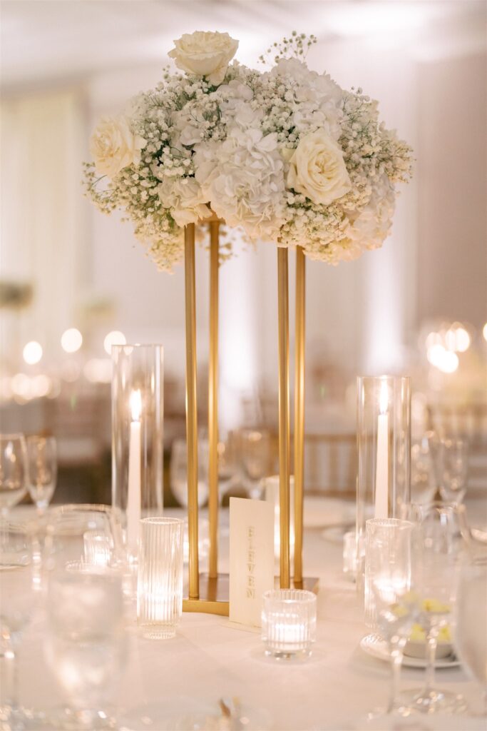 Tall gold centerpiece with white roses, hydrangeas, and baby's breath, surrounded by glass candles and table number on an elegant, softly lit table.