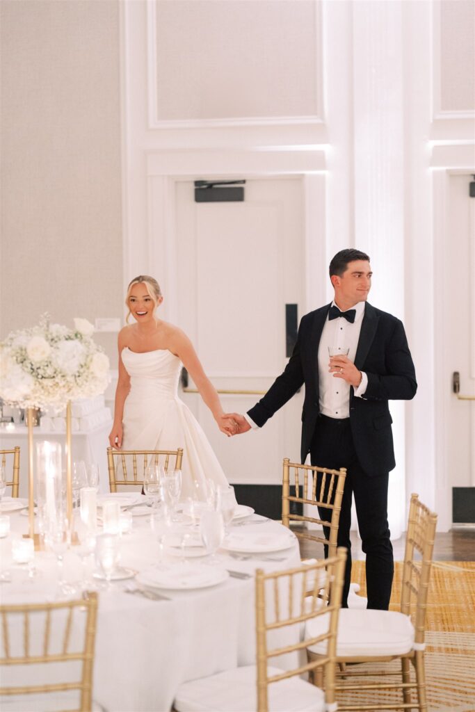 A bride and groom, dressed in formal wedding attire, hold hands while entering a decorated reception hall with round tables and gold chairs.