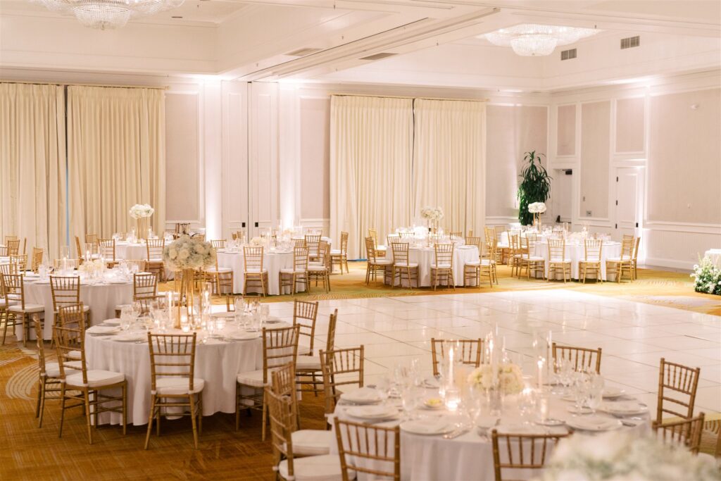 Elegant banquet hall with round tables set for an event, featuring white linens, gold chairs, floral centerpieces, and a large empty dance floor.