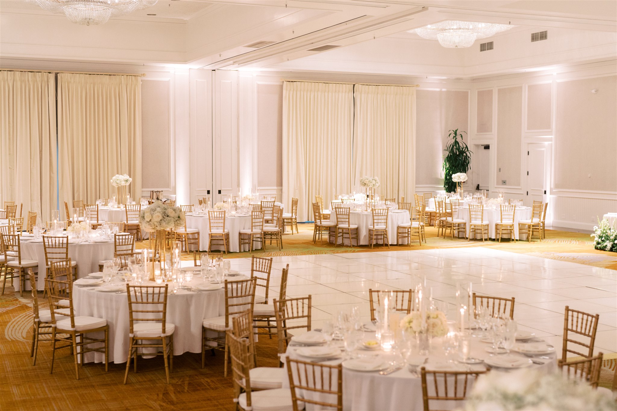 Elegant banquet hall with round tables set for an event, featuring white linens, gold chairs, floral centerpieces, and a large empty dance floor.