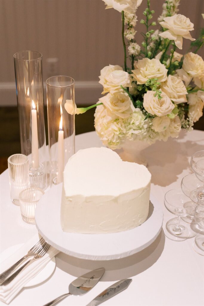 A heart-shaped white cake on a stand beside white flowers, lit candles in glass holders, stacked glasses, and tableware on a white tablecloth.