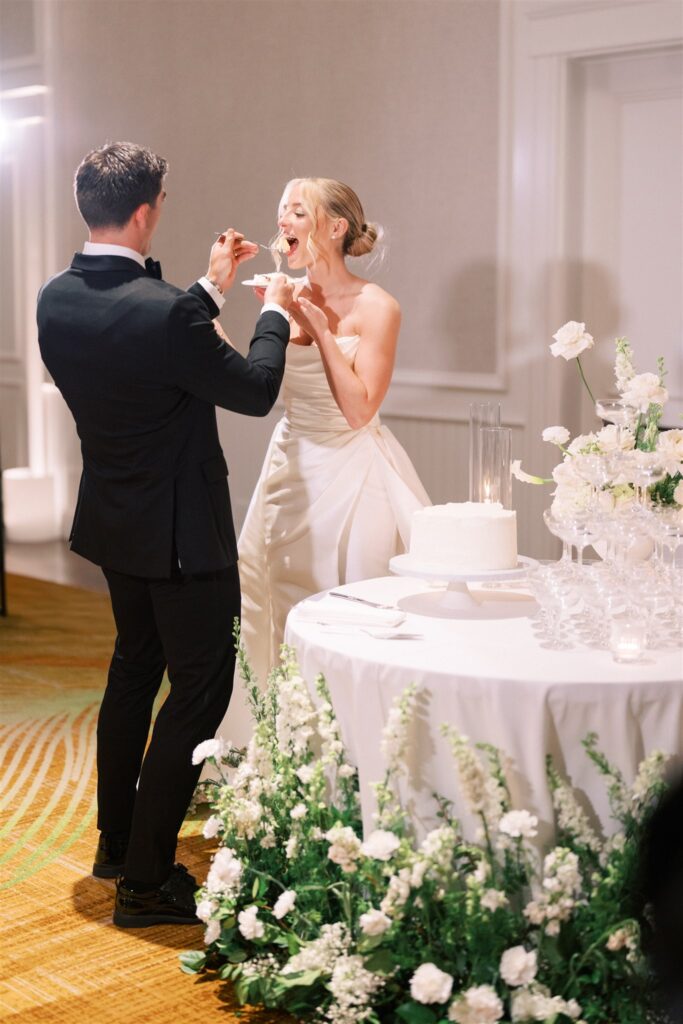 A bride and groom stand by a table with a wedding cake and flowers, feeding each other cake during their reception.