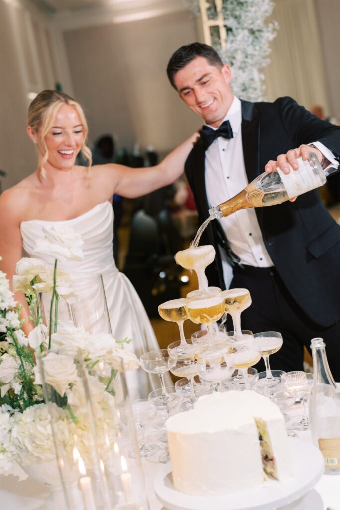 A woman in a white dress and a man in a tuxedo pour champagne into a tower of glasses at a decorated table with white flowers and a cake.