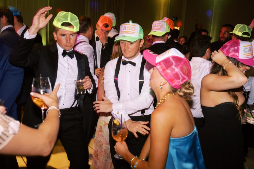 Several people dressed in formal attire and colorful neon hats dance and socialize at a lively indoor event or party.