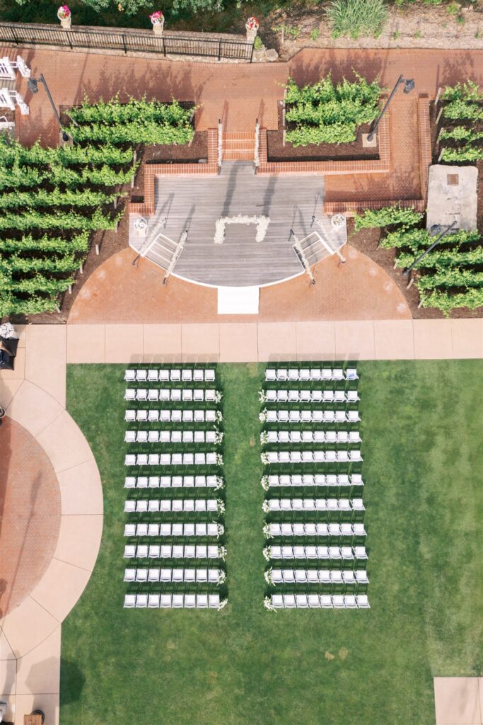 Aerial view of an outdoor wedding ceremony setup with rows of white chairs on grass facing a small stage with steps and brick accents.