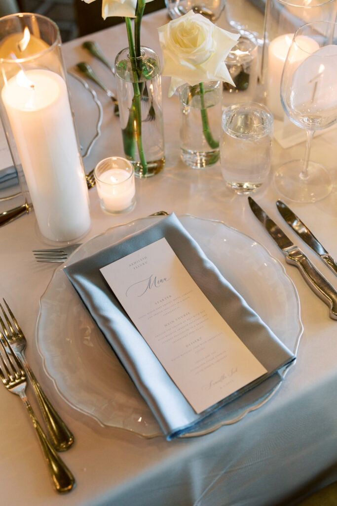 Elegant table setting with a menu on a light blue napkin atop a glass plate, surrounded by candles, white roses in vases, silver cutlery, and glassware.