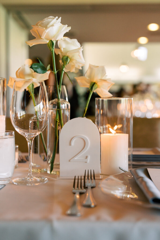 A table set for an event with white roses in vases, a glass of water, a wine glass, a lit candle, silverware, and a table number sign displaying the number 2.